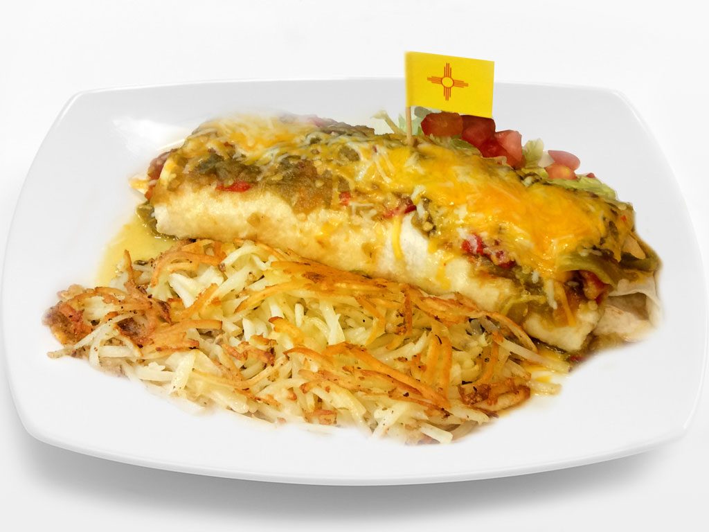 A burrito smothered in green chile and melted cheddar beside crispy hash browns, with a small New Mexico flag